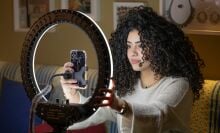 A woman with big, curly hair sits in front of her iPhone and a large ring light.