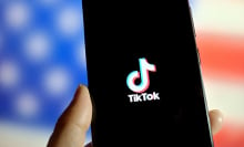 Person holds a phone showing the TikTok logo in front of a blurry American flag.
