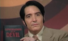 A man in a suit holds up a book while looking at the camera. The title reads "Conversations with the Devil".