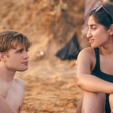 A young man and young woman sit on the beach looking romantically at each other.