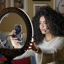 A woman with big, curly hair sits in front of her iPhone and a large ring light.