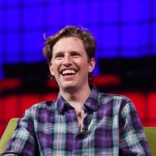 Matt Mullenweg is a white man with wavy blonde hair. He is wearing a plaid shirt and smiling on a lime green couch.