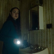 A woman in a dingy bathroom shines a torch off-camera, while looking afraid.