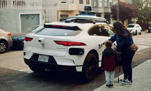 A woman and her child stand outside a white Waymo driverless vehicle, seen from the back. The woman's hand is on the car door's handle and it appears she is about to open the door.