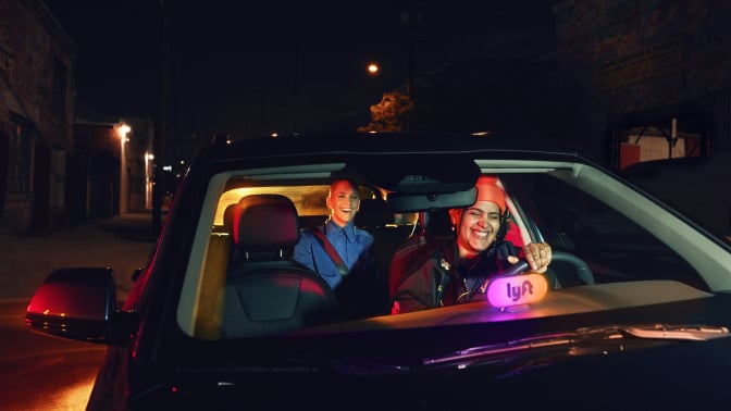 A person driving a Lyft smiles; a happy passenger rides in the back.