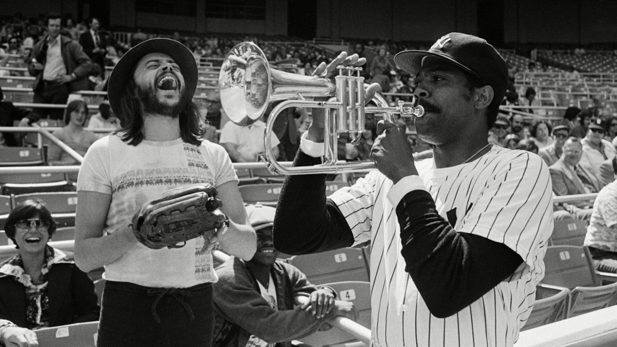Dock Ellis, wearing a New York Yankees uniform, plays a trumpet while jazz musician Chuck Mangione stands nearby, laughing.