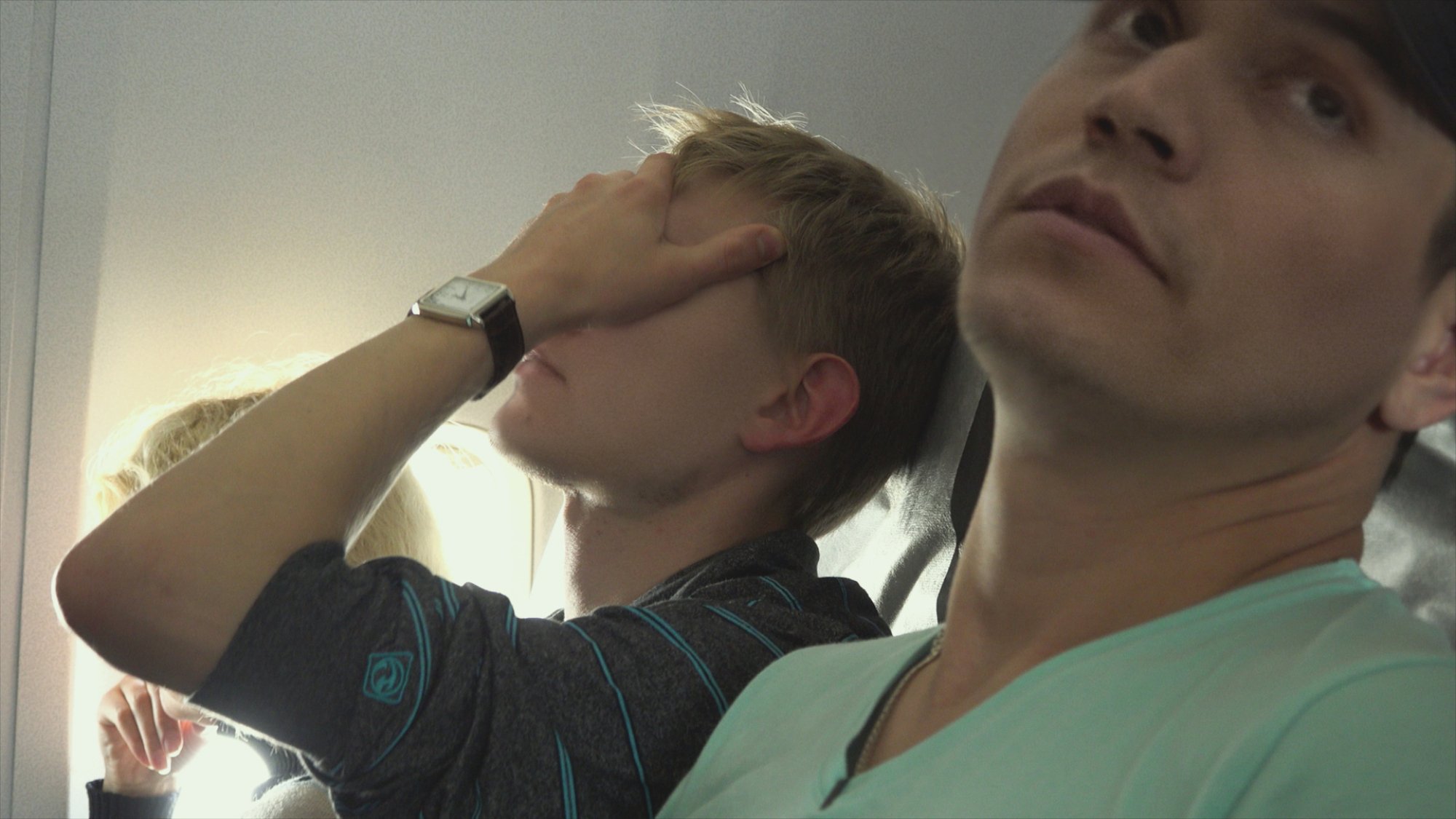 Two men seated on an airplane; one covers his face in worry.