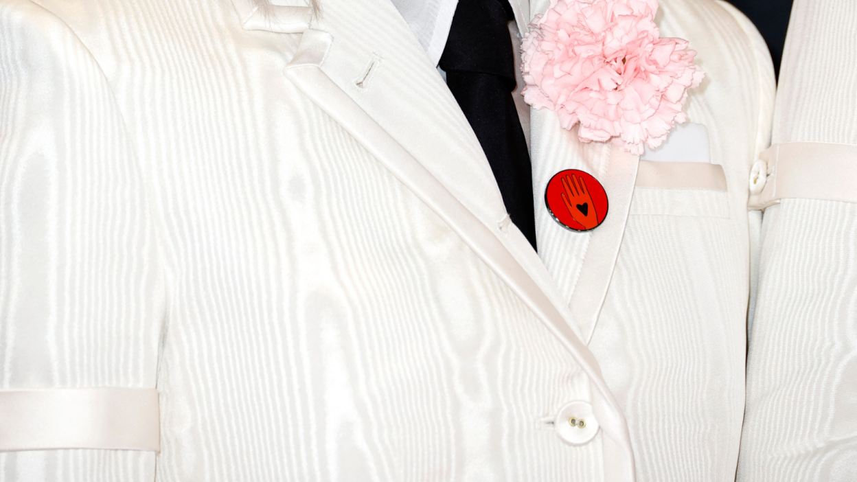 A zoomed-in photo of Phoebe Bridgers' white suit lapel, with a pink flower and a red "ceasefire now" button pinned to it.