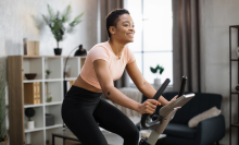 Smiling beautiful african american sports woman in sportswear cycling bike at home on background of light living room. Cardio training, exercising legs, cardio workout indoors.