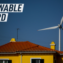 A picture shows the top of a typical portuguese house, yellow walls, dark green shutters, and a red rooftop. Behind it, against the blue sky a win turbine is peeking in the background.