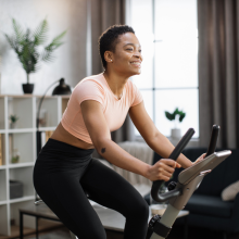 Smiling beautiful african american sports woman in sportswear cycling bike at home on background of light living room. Cardio training, exercising legs, cardio workout indoors.