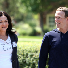 Sheryl Sandberg in a white graphic tee and navy blazer smiles and walks beside Zuckerberg in a navy pullover.