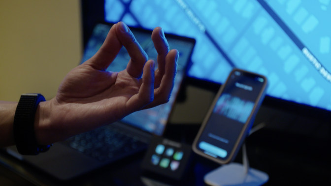 Close up of wrist shows a user wearing the Mudra while pinching their fingers. In the background, connected devices show the neural activity triggered by the hand gesture.